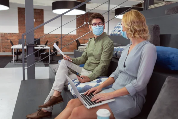 戴口罩的男女同事坐在大厅里讨论和使用平板电脑和笔记本电脑 在眼镜蛇大流行期间 在一家现代化的办公室工作 — 图库照片