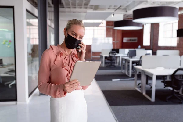 タブレットを使用してオフィスに顔マスクを着用し スマートフォンで話す白人実業家 コロナウイルスの流行期に近代的なオフィスで仕事をしていました — ストック写真