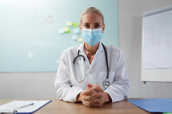 Kaukasische Ärztin Mit Gesichtsmaske Sitzt Schreibtisch Büro Und Hört Während — Stockfoto