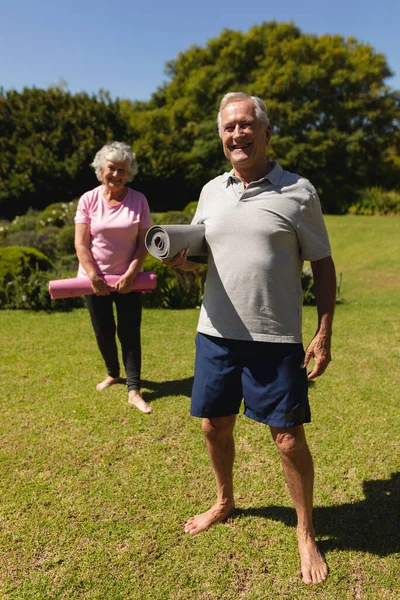 ヨガマットを持っているシニア白人夫婦の肖像画 カメラを見て 日当たりの良い庭で笑顔 定年退職と積極的なシニアライフスタイルのコンセプト — ストック写真