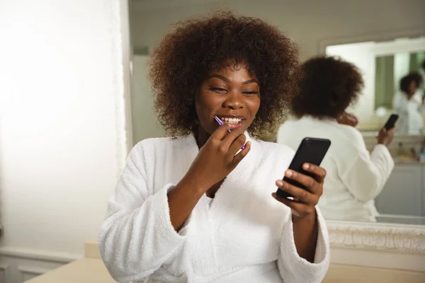 バスルームでアフリカ系アメリカ人女性の笑顔バスローブを着て 歯をブラッシングし スマートフォンを使用してください 健康美健康家庭で質の高い時間を過ごすこと — ストック写真
