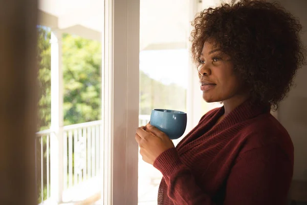 Счастливая Африканская Американка Выглядывающая Солнечного Окна Держащая Чашку Кофе Улыбающаяся — стоковое фото