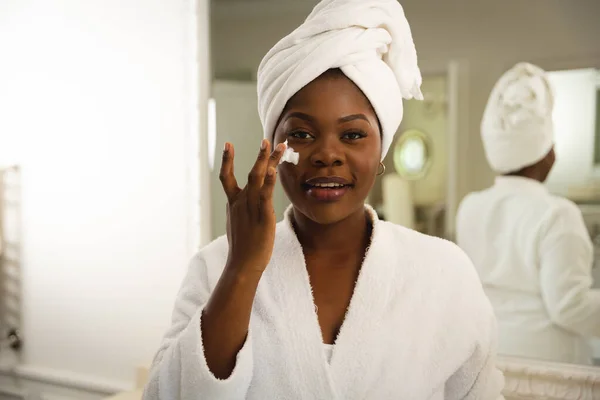 在浴室里笑着的非洲裔美国女人 头上拿着毛巾 看着镜子里湿透的脸 美丽和幸福 在家里度过高质量的时光 — 图库照片