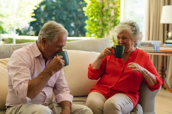 シニアの白人夫婦がリビングルームでコーヒーを飲みながらソファに座っています 隠遁と退職と幸せなシニアライフスタイルのコンセプト — ストック写真