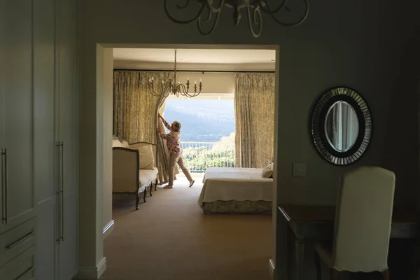 寝室の窓の拡大カーテンの隣に立つシニア白人女性 隠遁と退職と幸せなシニアライフスタイルのコンセプト — ストック写真