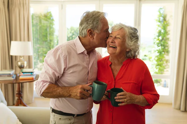 Senior Pareja Caucásica Besándose Mejilla Sonriendo Sosteniendo Tazas Salón Retiro —  Fotos de Stock