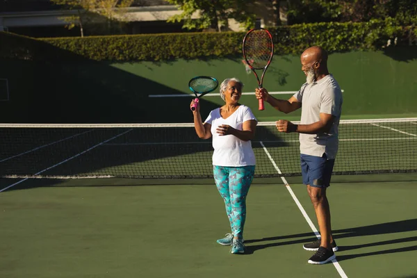 在网球场上挂着网球拍的笑着的资深非洲裔美国夫妇的画像 退休和积极的老年生活方式概念 — 图库照片