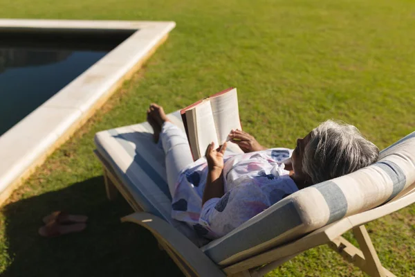 日当たりの良い庭でプールでデッキチェアで本を読んでいるシニアアフリカ系アメリカ人女性 隠遁と退職と幸せなシニアライフスタイルのコンセプト — ストック写真