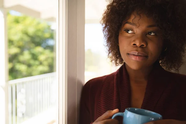 富有洞察力的非洲裔美国女人举着一杯咖啡从阳光灿烂的窗户往外看 在家里消磨时间 — 图库照片