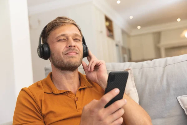 Homem Caucasiano Sentado Sofá Usando Fones Ouvido Usando Smartphone Casa — Fotografia de Stock