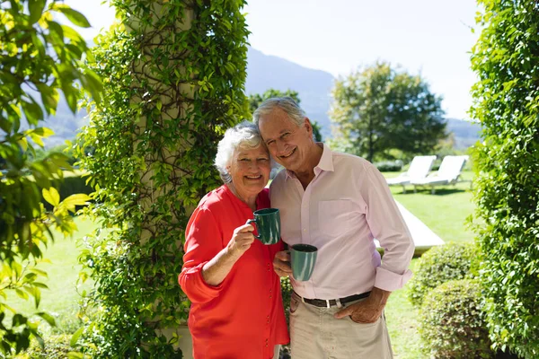 日当たりの良い庭で シニア白人夫婦の笑顔と保持マグカップの肖像画 隠遁と退職と幸せなシニアライフスタイルのコンセプト — ストック写真