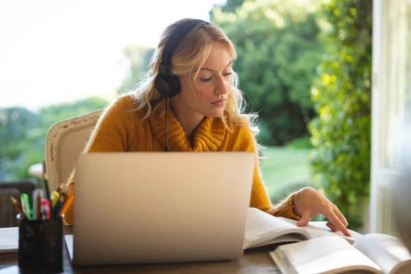 自宅のリビングルームで働き ヘッドフォンを着用し ノートパソコンを使用し 本を読む白人女性 柔軟性のあるテクノロジーを使って — ストック写真