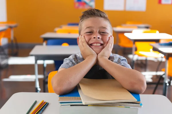 教室の机の上に座って本にもたれて笑っている幸せな白人の学童 子供の頃と小学校での教育は — ストック写真