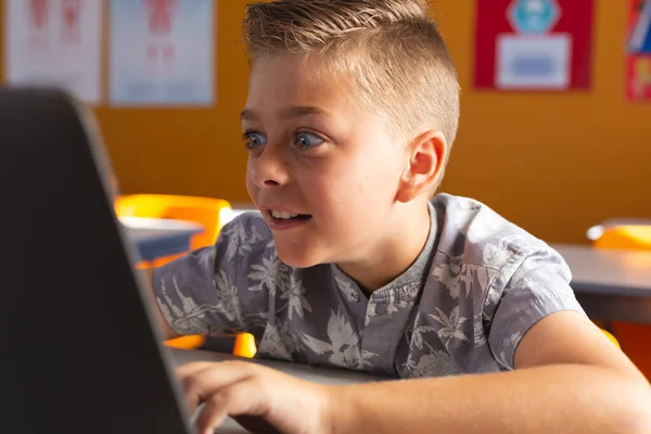兴奋的高加索男孩坐在课桌前 用笔记本电脑笑着 小学的儿童 技术和教育 — 图库照片