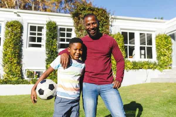 一个笑容满面的非洲裔美国父亲的画像 他带着儿子在阳光灿烂的花园里拥抱和举行足球 家庭呆在家里的时间 — 图库照片