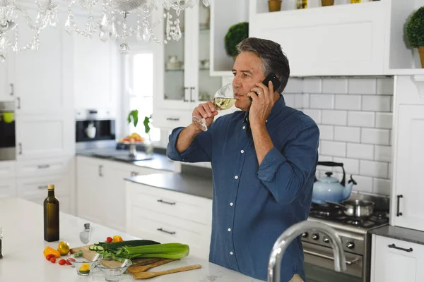 在现代厨房里 一位高年级的高加索人 一边讲着智能手机 一边喝酒 退休生活方式 呆在家里 — 图库照片