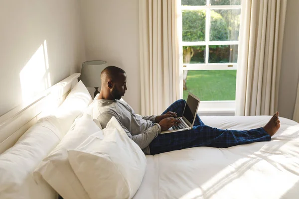 アフリカ系アメリカ人の男性がノートパソコンを使ってベッドの上に寝そべっている テクノロジーで家で一人で過ごすのは — ストック写真