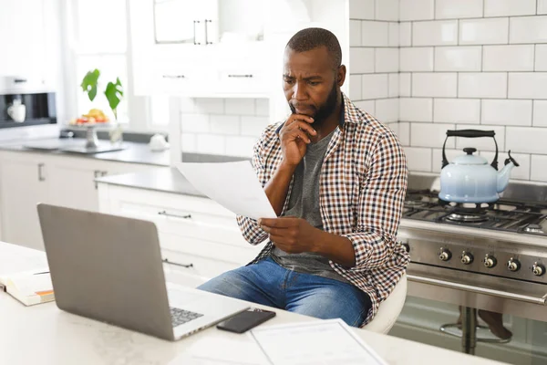 Poważny Afrykański Amerykanin Siedzący Kuchni Przeglądający Papiery Korzystający Laptopa Zdalna — Zdjęcie stockowe