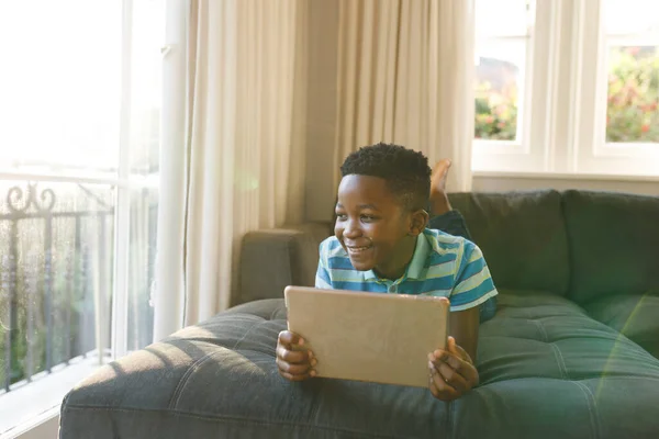 アフリカ系アメリカ人の男の子がタブレットを使って笑顔でリビングでソファに横になっています テクノロジーだけで家で過ごす時間は — ストック写真