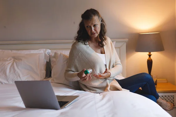 卧室里的高个子高加索女人坐在床上 拿着一盒药丸 用笔记本电脑 退休的生活方式 独自呆在家里用科技 — 图库照片