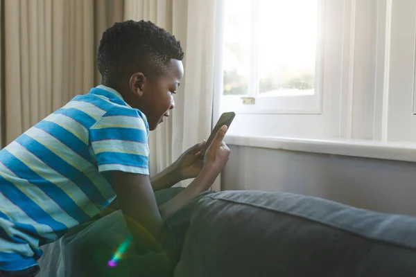 Χαρούμενο Αφροαμερικάνικο Αγόρι Γονατίζει Στον Καναπέ Χρησιμοποιώντας Smartphone Στο Σαλόνι — Φωτογραφία Αρχείου