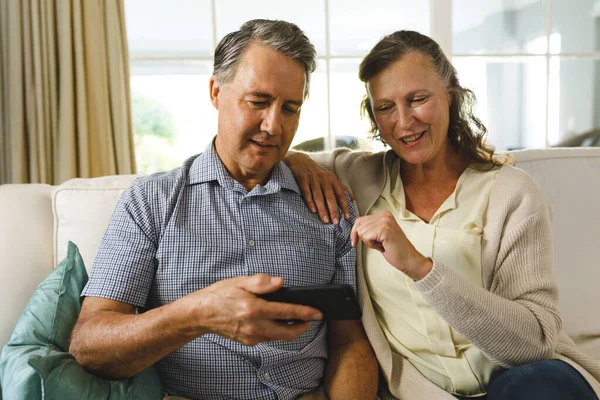 リビングで幸せな高齢者の白人カップルは ソファに座って スマートフォンを使用しています 引退後の生活や家で過ごす時間 — ストック写真