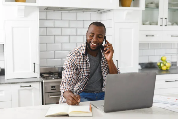 Uśmiechnięty Afrykanin Siedzący Kuchni Pracujący Laptopie Smartfonie Zdalna Praca Domu — Zdjęcie stockowe