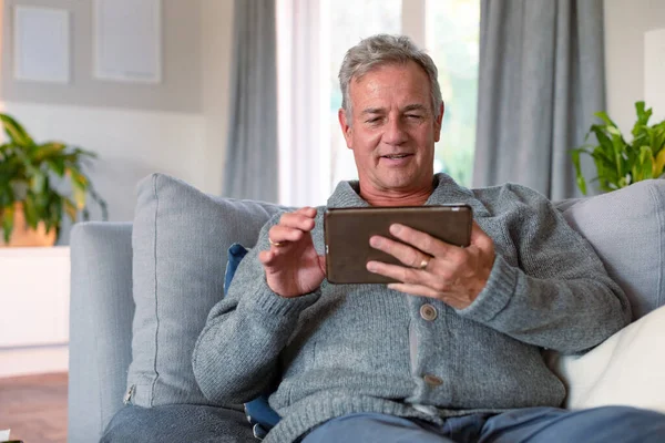 幸せな白人の先輩がソファに座ってタブレットを使っています 家庭での積極的な退職生活 — ストック写真