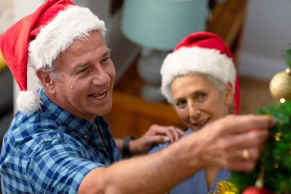 サンタの帽子をかぶった幸せな白人のシニアカップルは クリスマスツリーを飾る 家庭でのクリスマスやお祭りや伝統 — ストック写真