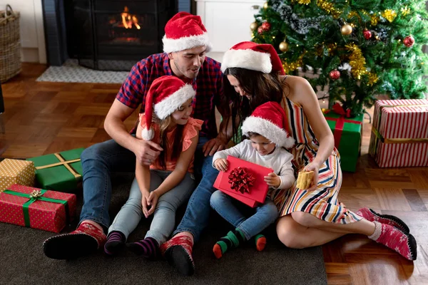 サンタの帽子をかぶった幸せな白人の家族は クリスマスの時にプレゼントを持っています クリスマスの時間と祭りを一緒に家で — ストック写真