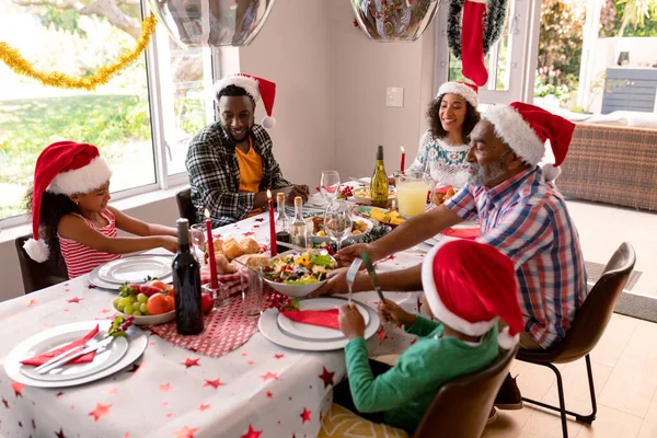 サンタの帽子をかぶった幸せなマルチ世代の家族は クリスマスの食事をしています クリスマスの時間と祭りを一緒に家で — ストック写真