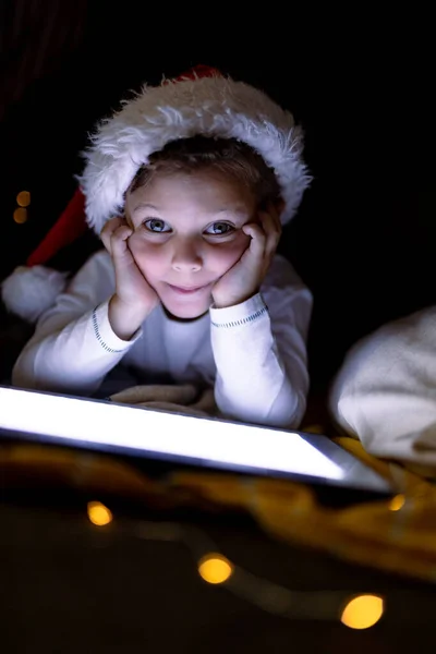 白人の少年サンタの帽子をかぶって タブレットを使用して クリスマスの時間にカメラを見て 家庭での子供時代やクリスマスやお祭りや伝統 — ストック写真