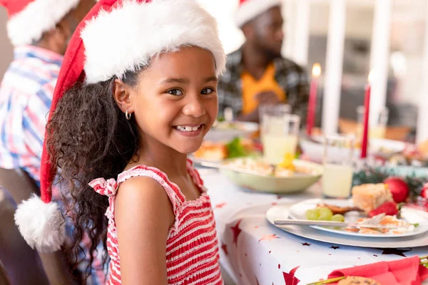 クリスマスのテーブルに座ってサンタの帽子をかぶった幸せなアフリカ系アメリカ人の少女 クリスマスの時間と祭りを一緒に家で — ストック写真