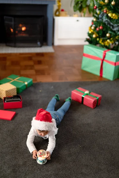 Focused Caucasian Boy Wearing Santa Hat Looking Snow Globe Christmas — Stock Photo, Image