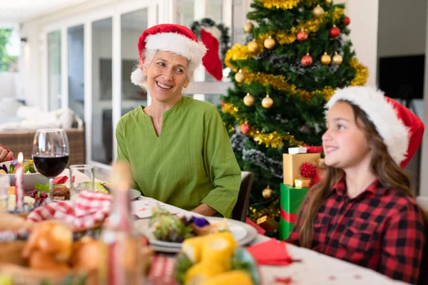 Vrolijke Blanke Grootmoeder Kleindochter Met Kerstmutsen Die Aan Kersttafel Praten — Stockfoto