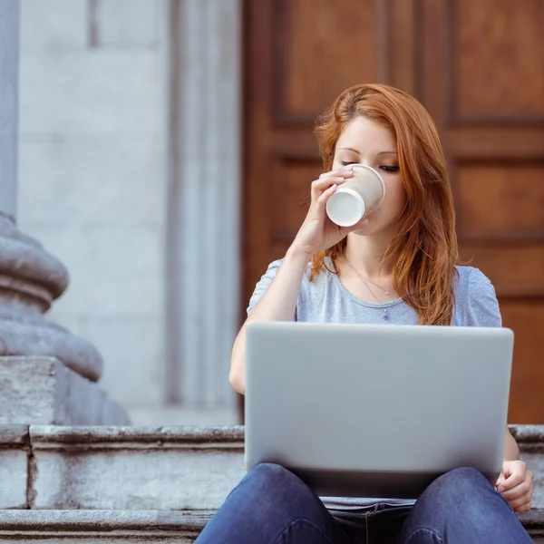 Hermosa mujer bebiendo de taza desechable usando su computadora portátil — Foto de Stock