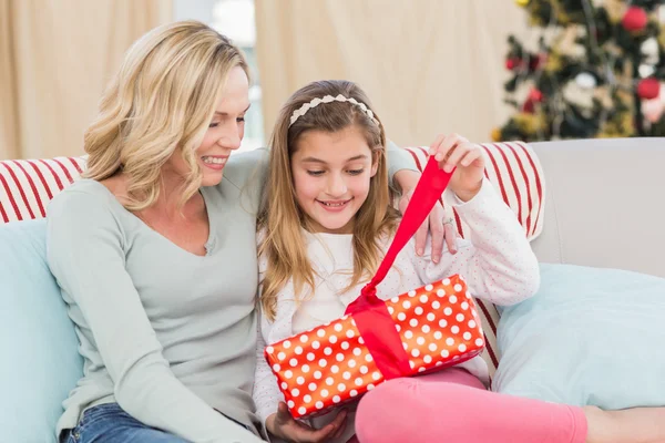 Cute little girl sitting on couch opening gift with mum — Stock Photo, Image