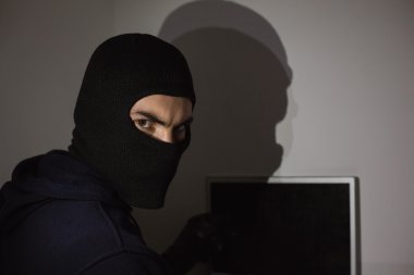 Hacker in balaclava hacking laptop while looking at camera clipart