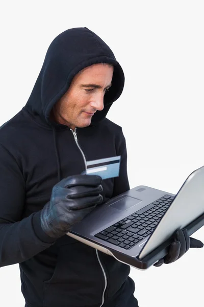 Smiling thief in hood jacket using laptop and credit card — Stock Photo, Image