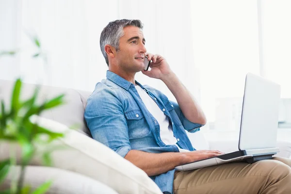 Homem alegre sentado no sofá telefonando e usando laptop — Fotografia de Stock