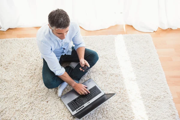 Hombre sentado en la alfombra usando el ordenador portátil y el teléfono — Foto de Stock