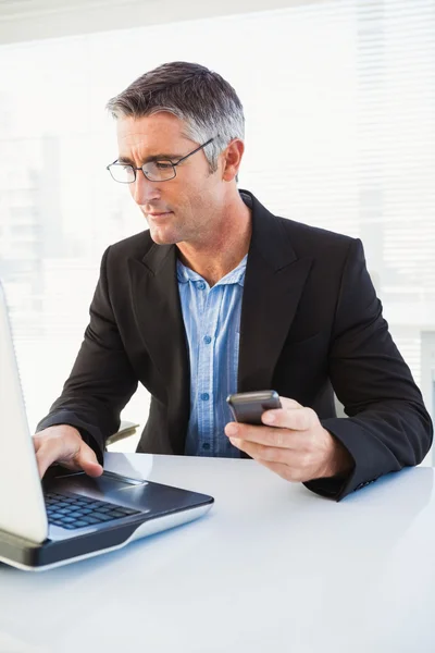 Uomo d'affari allegro utilizzando laptop e tenendo smartphone — Foto Stock