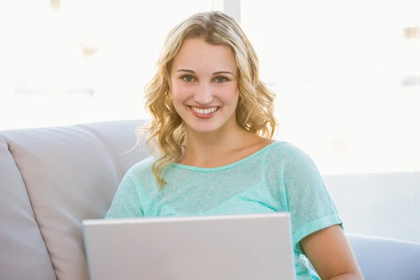 微笑的金发美女坐在沙发上使用笔记本电脑的肖像 图库图片