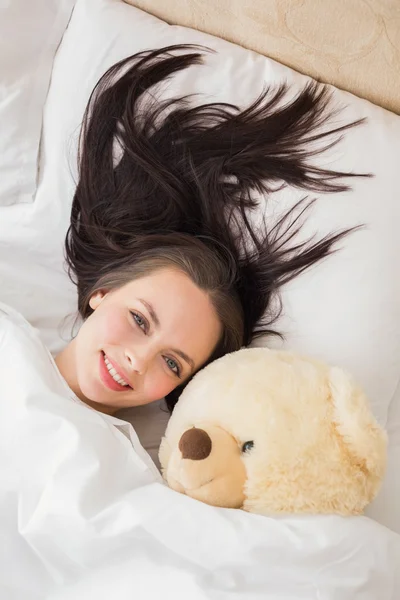 Hübsche Brünette unter der Bettdecke mit Teddybär — Stockfoto
