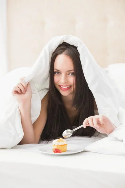 Pretty brunette eating cupcake in bed — Stock Photo, Image