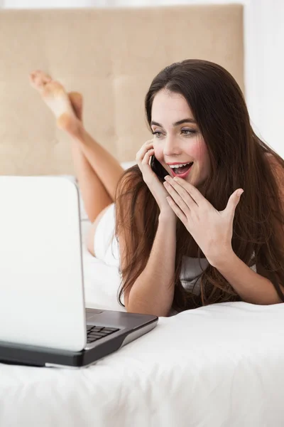 Söt brunett på telefonen i säng med laptop — Stockfoto