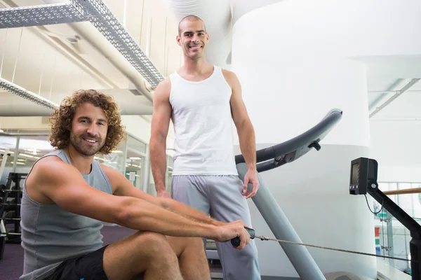 Allenatore maschile che assiste l'uomo su attrezzo fitness in palestra — Foto Stock