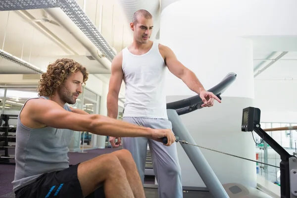 Allenatore maschile che assiste l'uomo su attrezzo fitness in palestra — Foto Stock