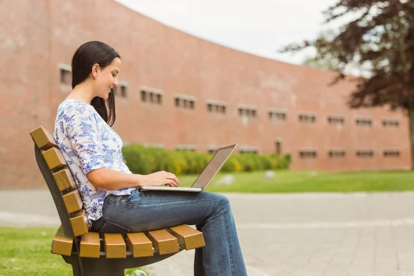 Glad brunett sitter på bänken att skriva på laptop — Stockfoto