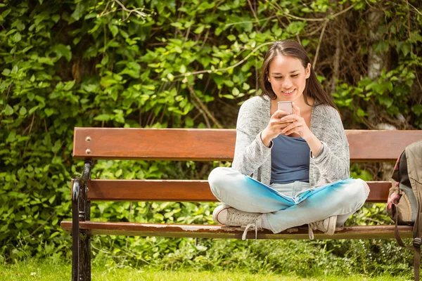Étudiante souriante assise sur un banc message texte sur son téléphone mobile — Photo